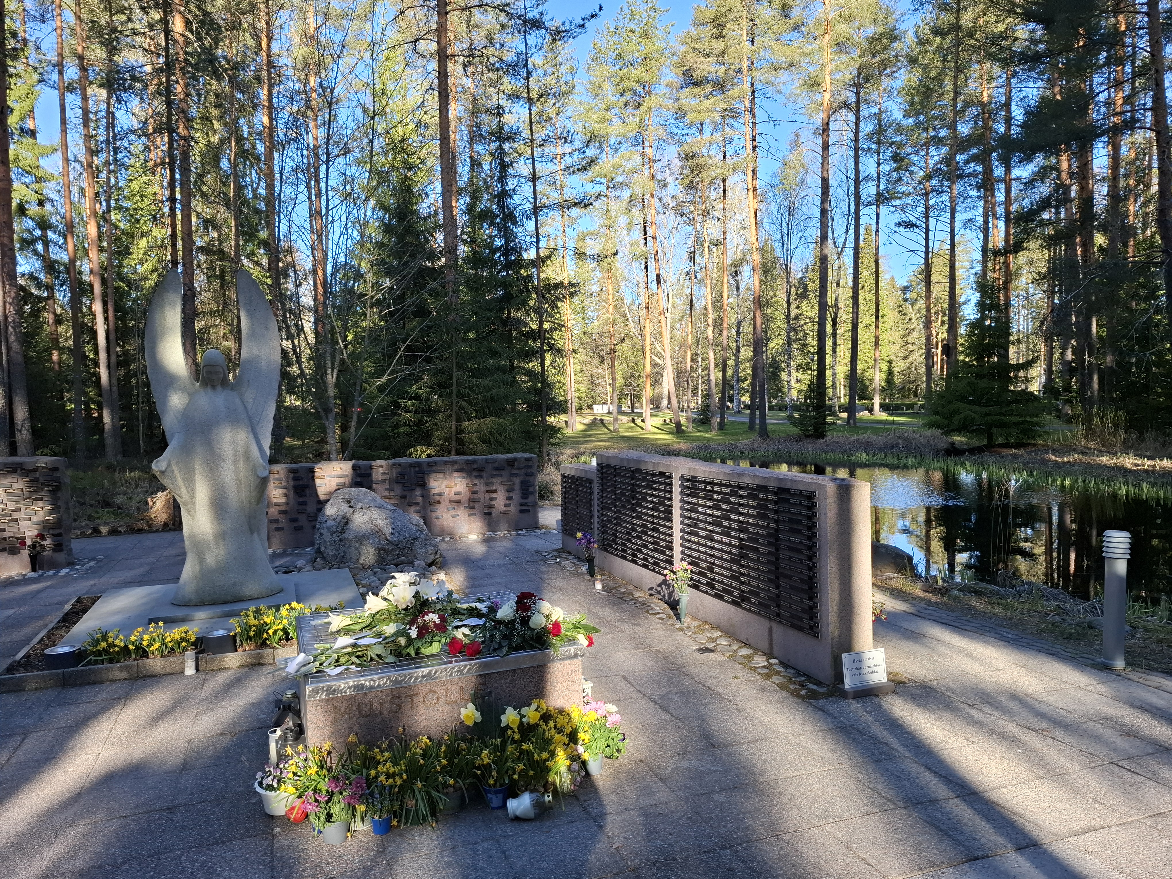 Hautausmaalla muistomerkki, enkelipatsas ja kukkia.