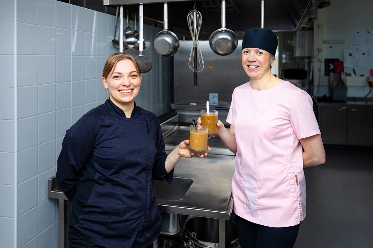 Kaksi keittiöammattilaista rosterisen työtason edessä käsissään jääkahvit hymyilevät kameralle.