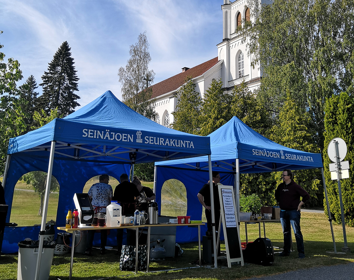 Seinäjoen seurakunnan kaksi sinistä paviljonkitelttaa joiden sisällä seurakunnan työntekijöitä, taustalla Y...