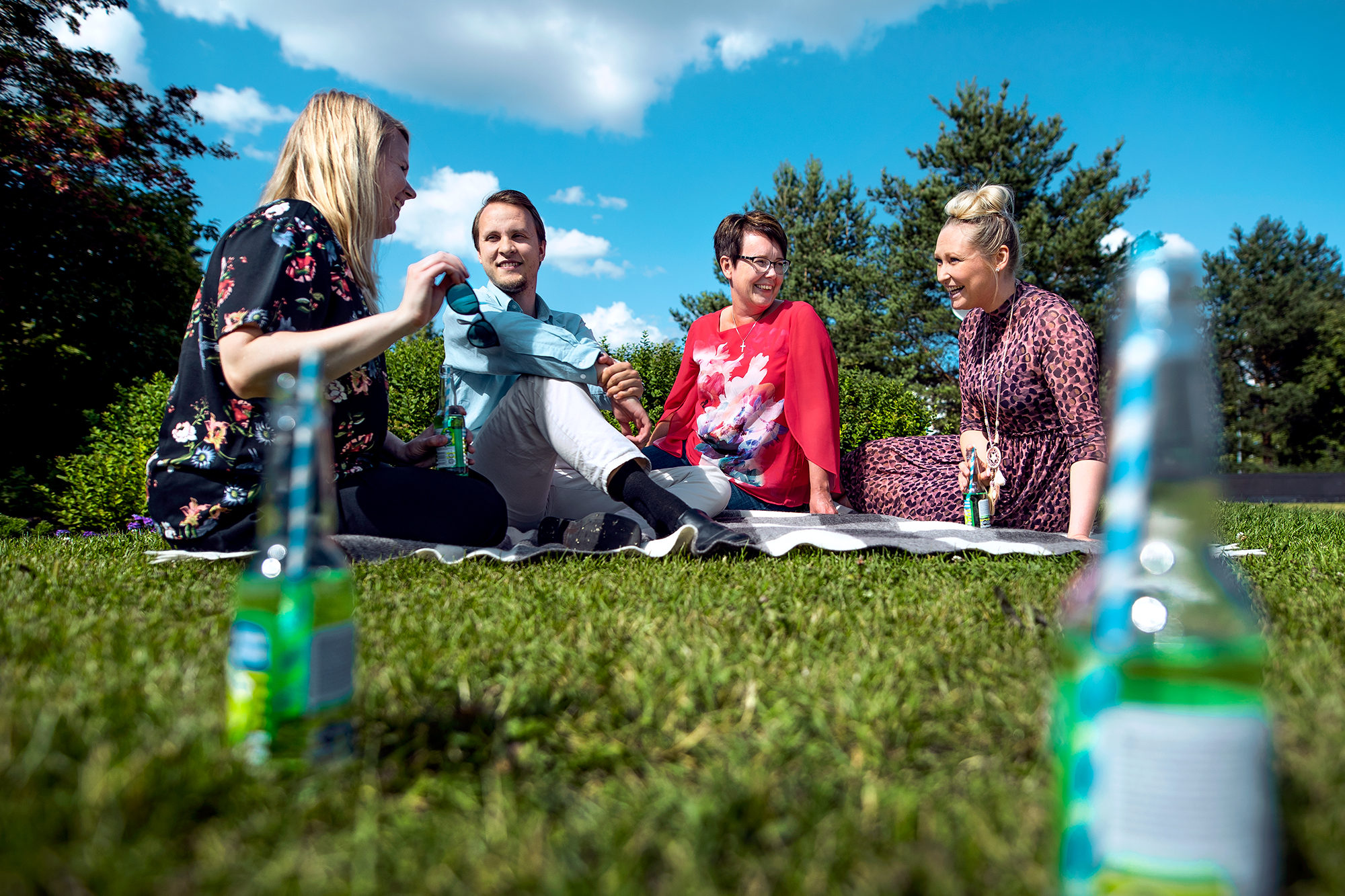Neljä ihmistä istuu nurmikolla viltillä nauraen ja juoden limonadia.