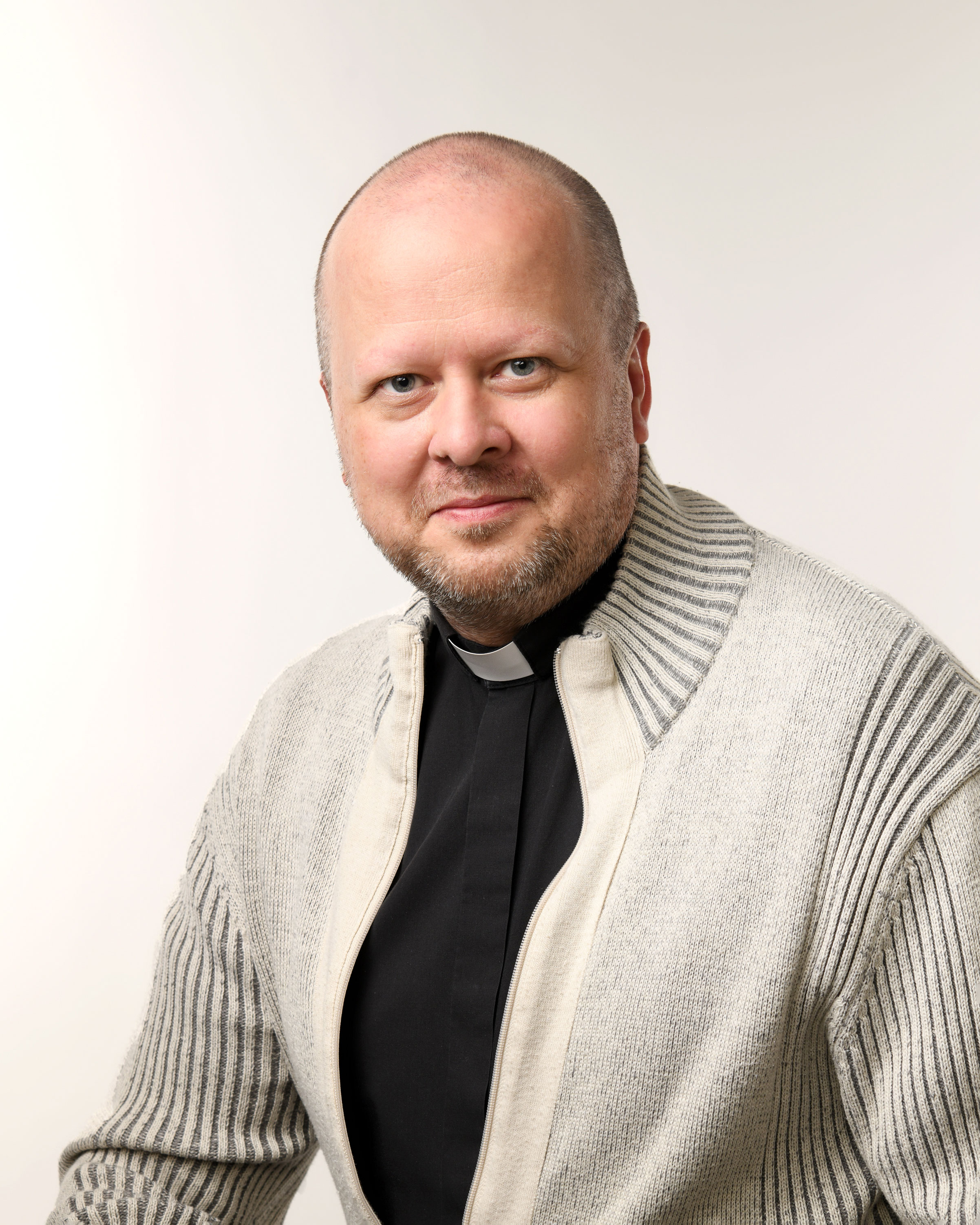 Teemu Leino katsoo kameraan. Hän on pukeutunut papin paitaan ja vaaleaan villatakkiin.