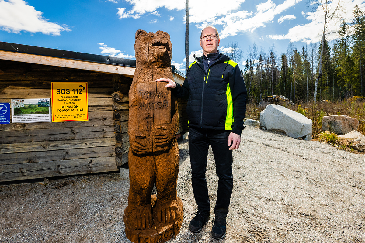 Puusta veistetty karhupatsas ja henkilö vierekkäin metsäisessä maisemassa.